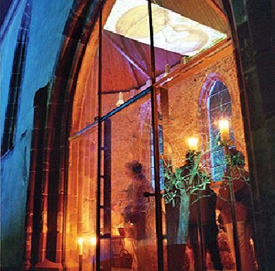 Beleuchteter Kapelleninnenraum bei einer Veranstaltung am Abend, durch eine Rundbogenglastür gesehen